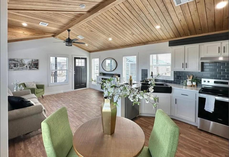 Modern living and kitchen area with wooden ceiling, light wood floors, green chairs, and floral decor. Bright, airy ambiance with white cabinets.