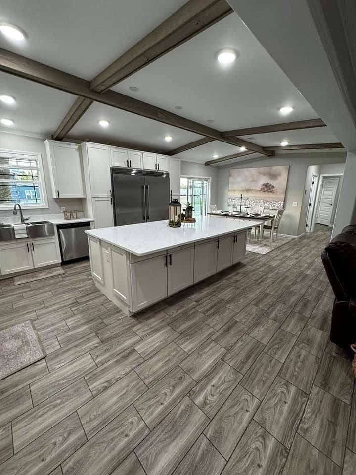 Modern kitchen with a white island and stainless steel appliances. Exposed wooden beams, tile flooring, and a dining area with artwork create a cozy ambiance.