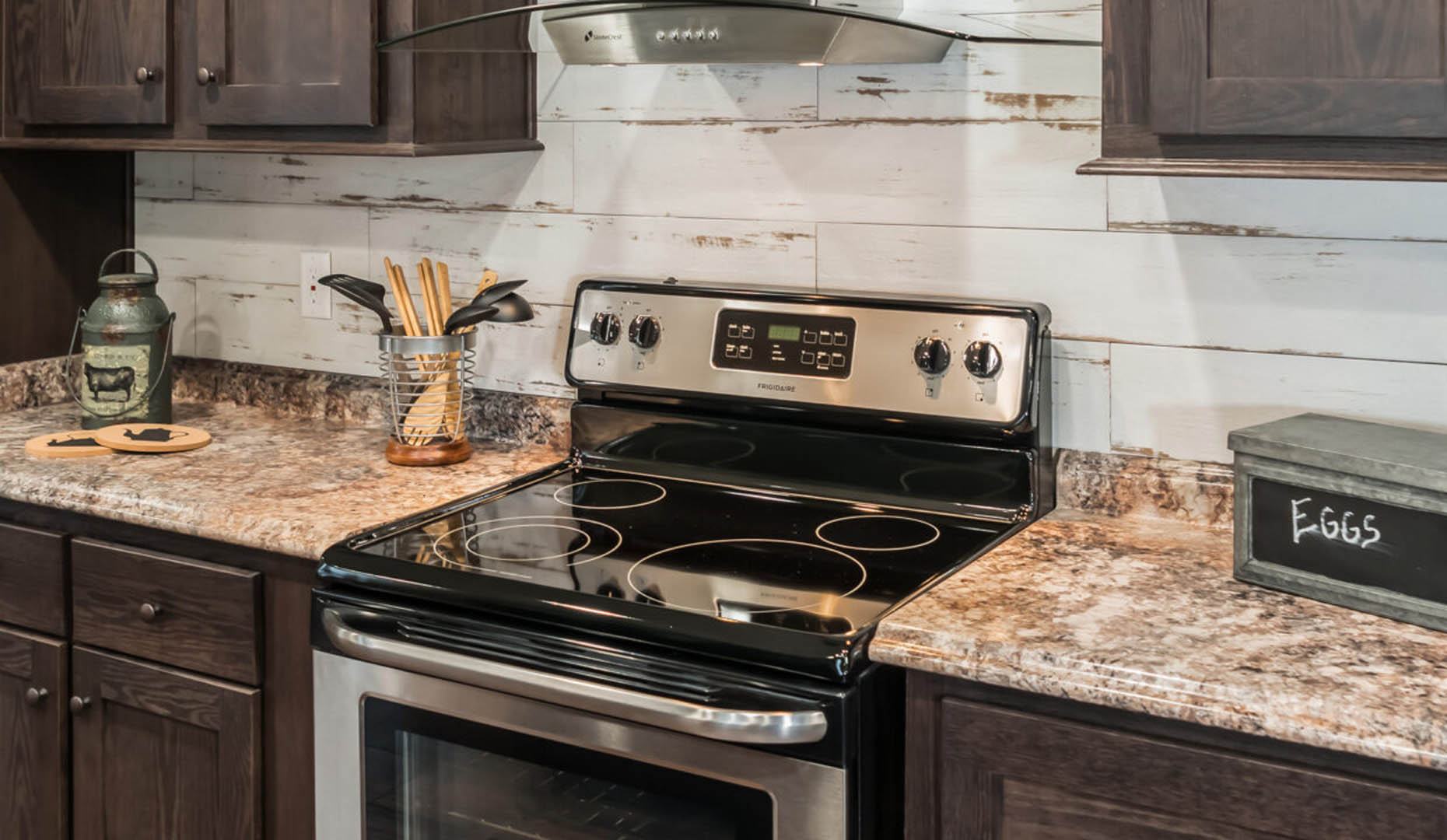 Modern kitchen with dark wood cabinets, marble countertops, and a stainless steel electric stove. Rustic decor includes an eggs box and utensils jar.