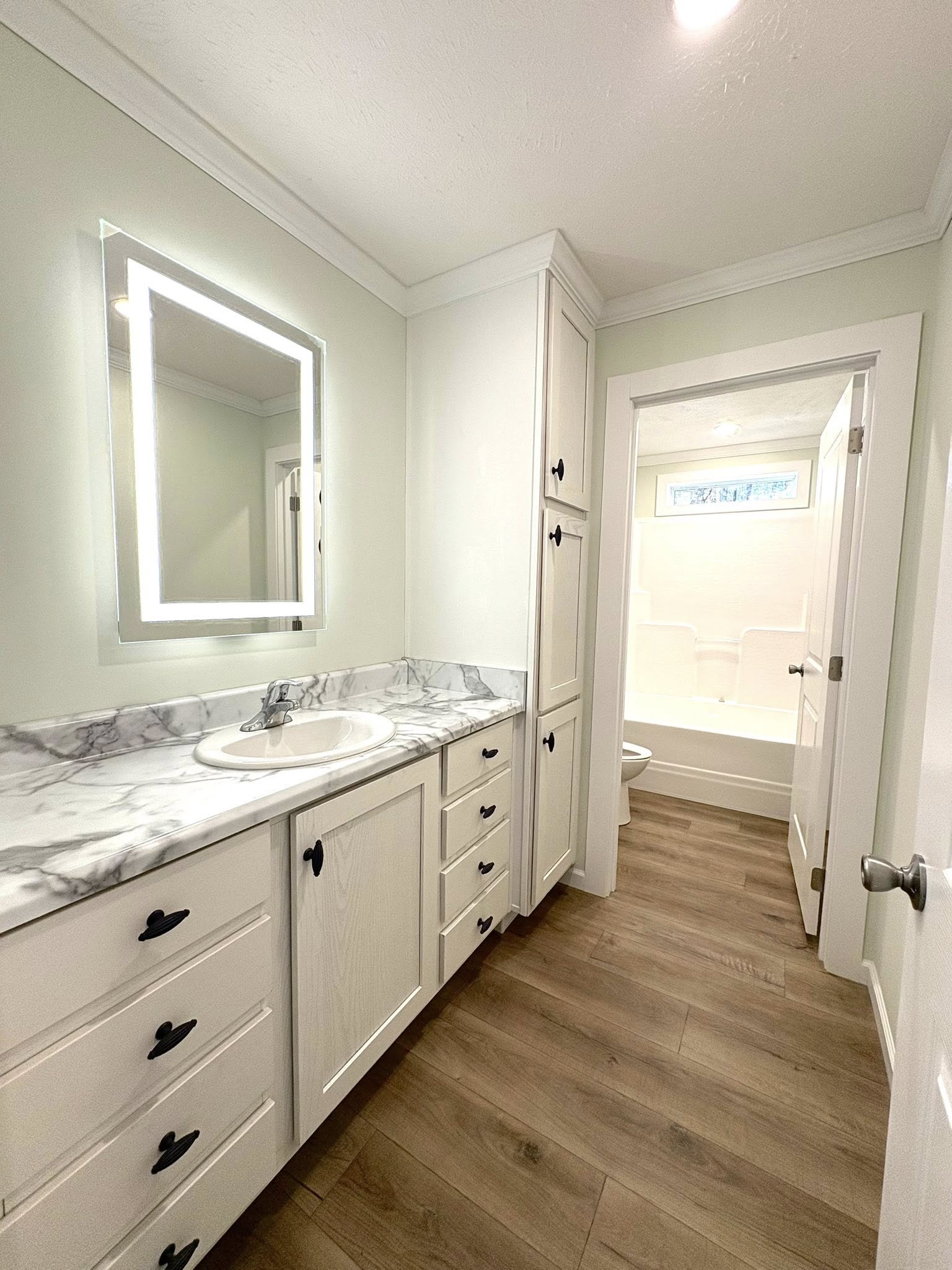 Bright bathroom with a marble countertop, white cabinets, and a backlit mirror. Wooden floor leads to a tub visible through the open door, creating a clean, modern feel.