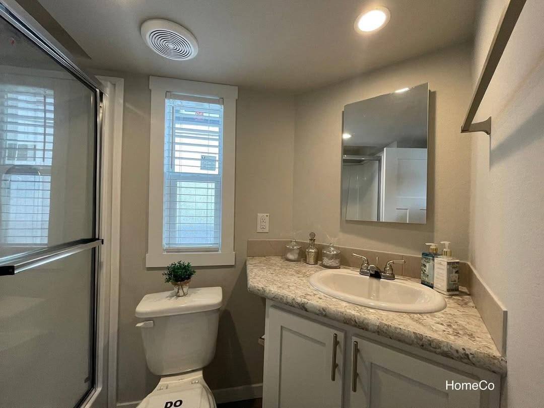 Modern bathroom with a marble countertop, under-mount sink, and rectangular mirror. Neutral tones, glass shower, and a small plant on the toilet.