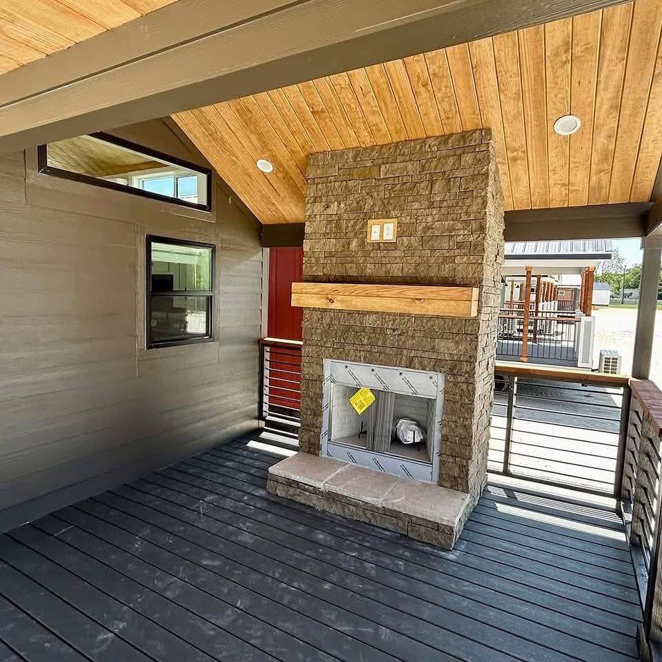 Covered patio with wooden ceiling and gray flooring, features a stone fireplace with a wooden mantel. A small window and exterior view add brightness.