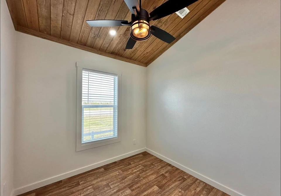 Small, empty room with warm wood ceiling and floor, white walls, a window with blinds, and a modern ceiling fan with a light, creating a cozy vibe.