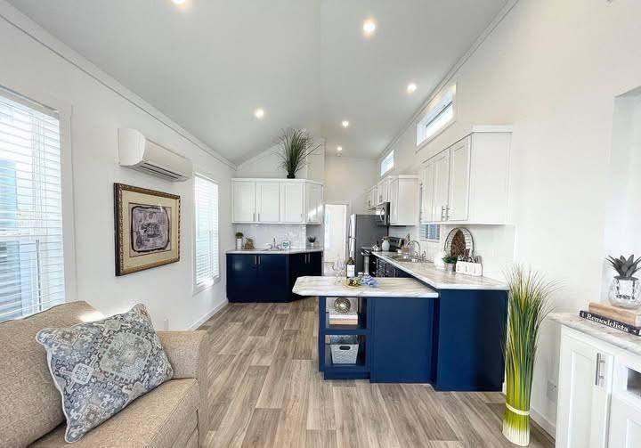 Bright, modern kitchen-living space with white walls, high ceiling, and spotlights. Navy lower cabinets, white upper cabinets, marble countertops, wood floor, decor with plants, and artwork create a cozy, inviting atmosphere.