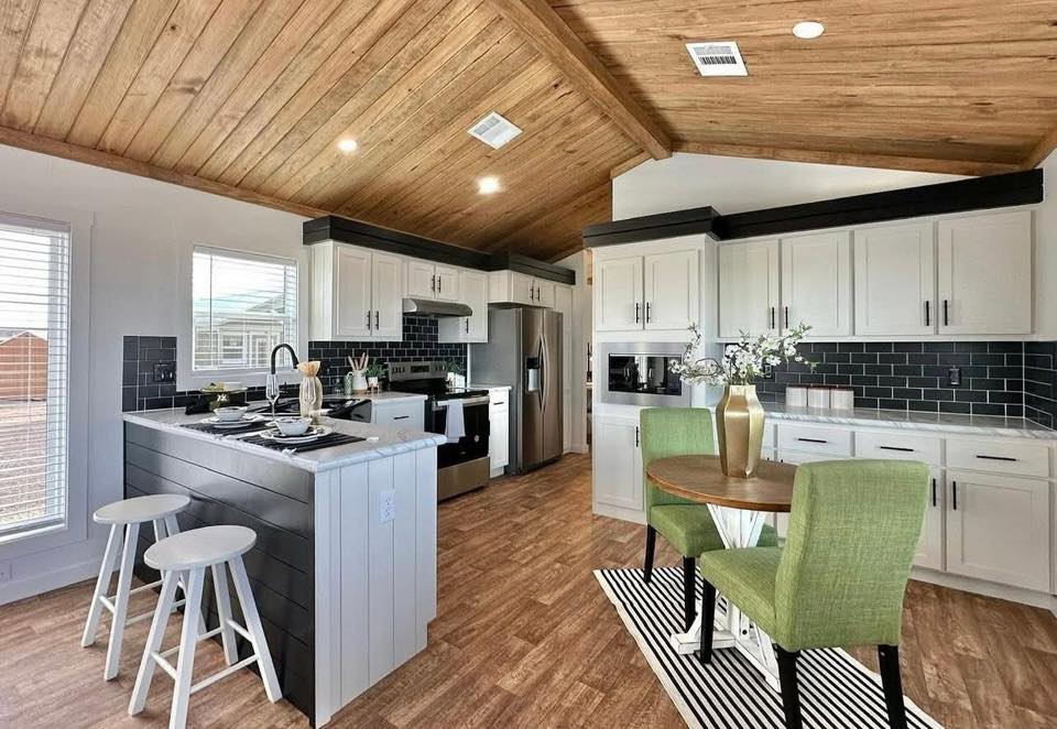 A modern kitchen with wooden ceilings, white cabinets, and black tile backsplash. Includes an island with stools and a small dining table with green chairs.