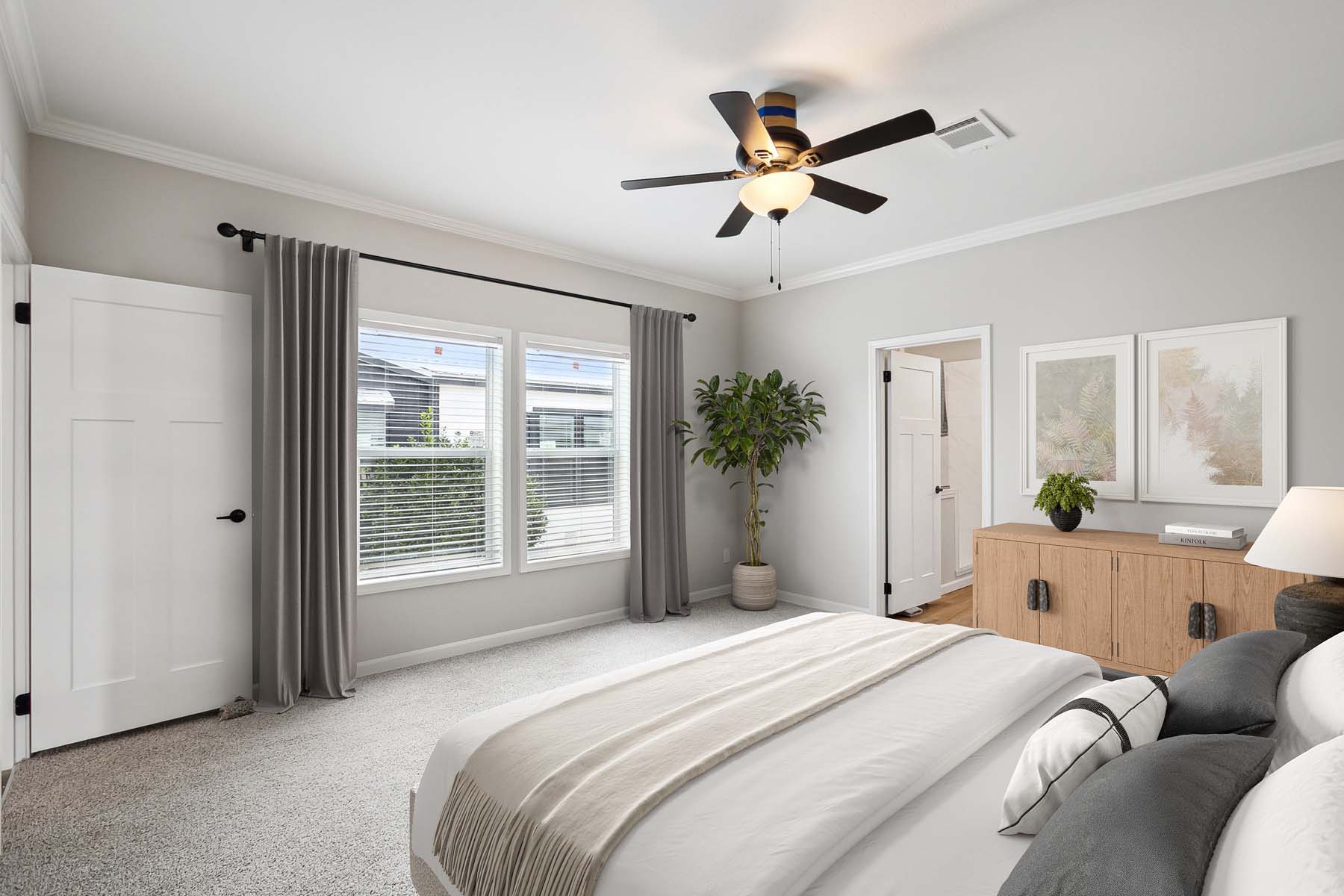 A serene bedroom with soft gray walls and carpet. Features a neatly made bed, large window with drapes, ceiling fan, potted plant, and wooden dresser. Neutral and calming atmosphere.