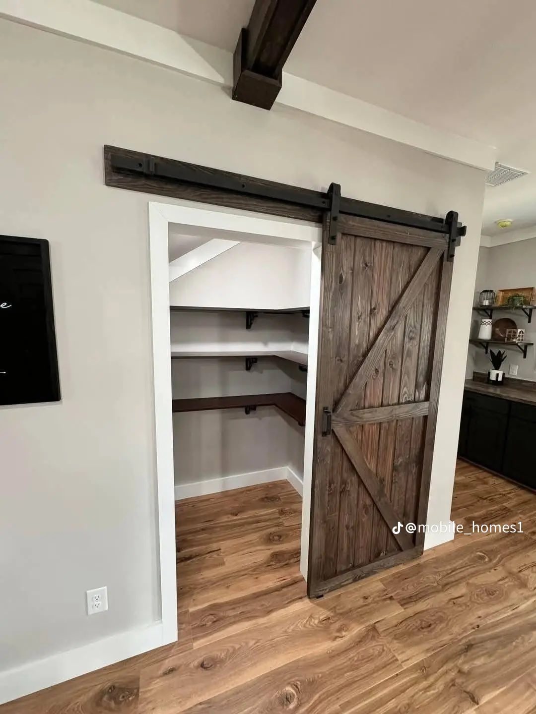 Rustic barn door slides open to reveal a small closet with corner shelves. The space features wood floors, a neutral wall, and a cozy, homely ambiance.