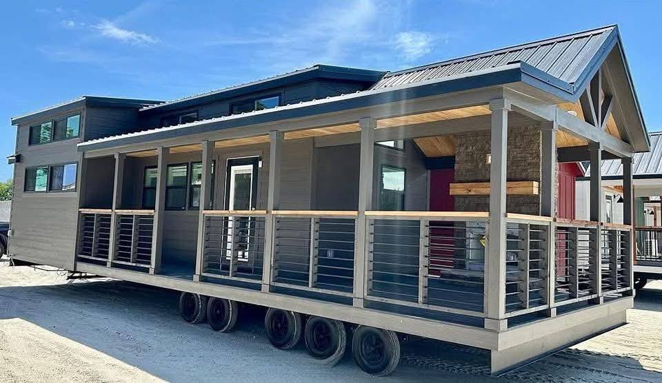 A modern tiny house on wheels with a covered porch and large windows, set against a clear blue sky. The design is sleek and inviting.