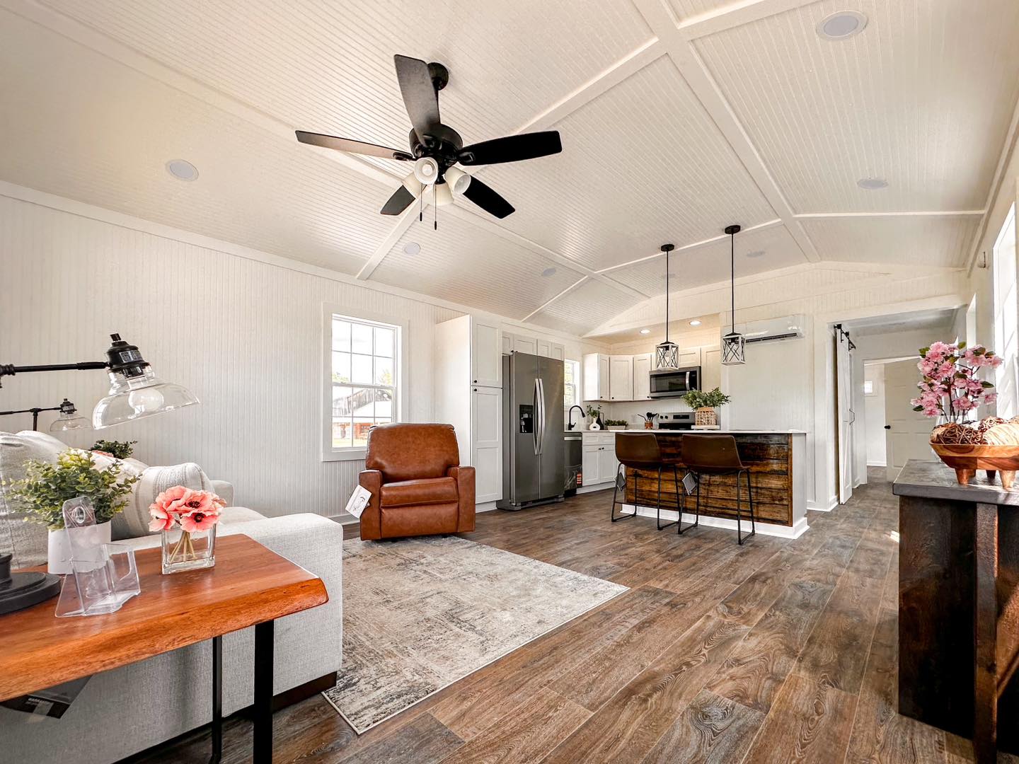 Spacious modern living room with a vaulted ceiling and wooden floors, featuring a brown recliner, rustic kitchen island, and ceiling fan, conveying warmth.