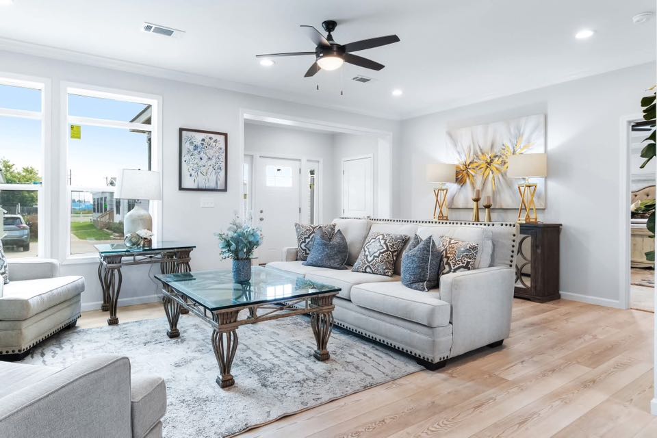 Elegant living room with light gray sofa, decorative pillows, and glass coffee table on a patterned rug. Bright, airy vibe with large windows.