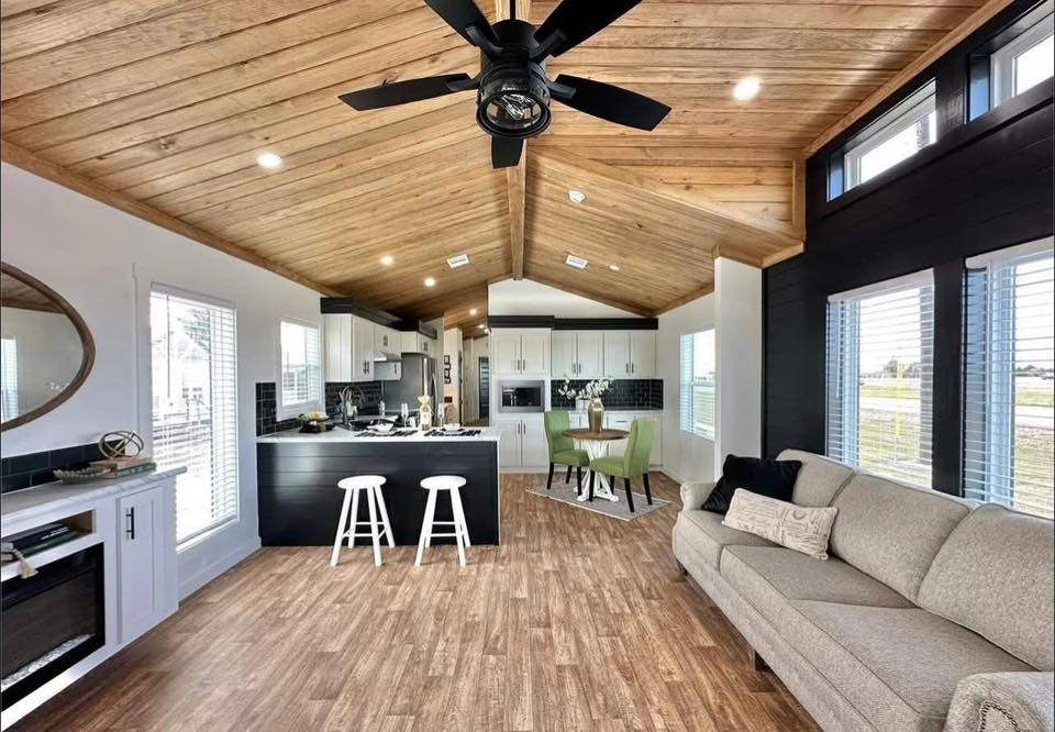 Spacious living area with a vaulted wooden ceiling, fan, and wood flooring. Features a gray sofa, kitchen with island and stools, and a dining nook.