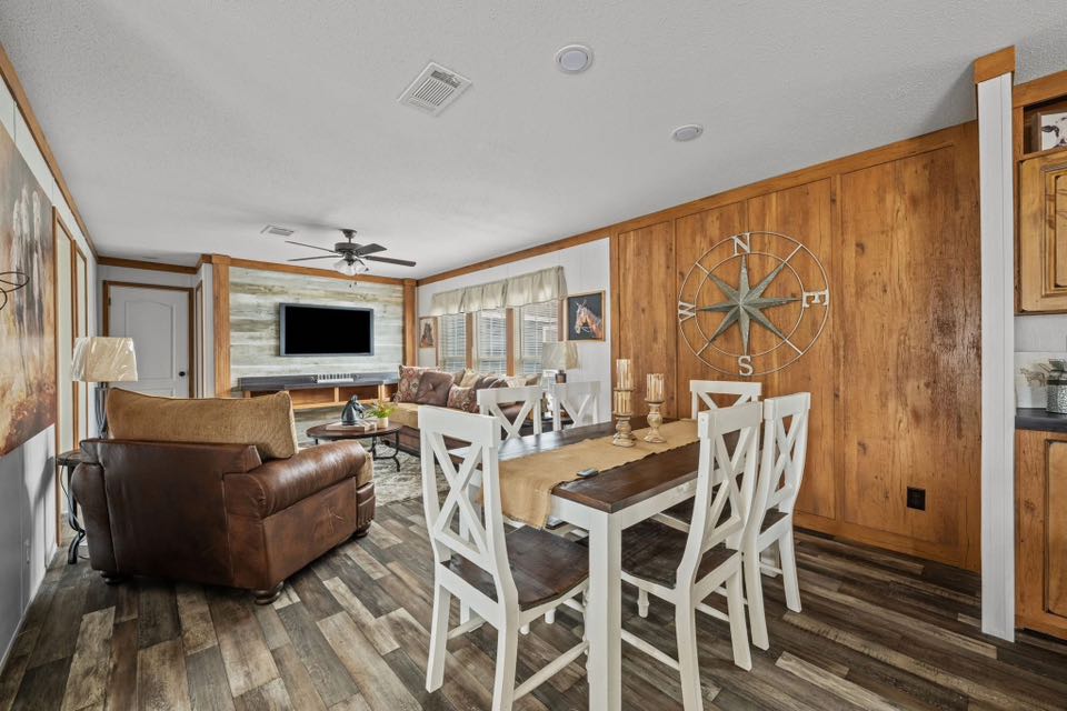 Cozy living-dining area with rustic wooden walls, a compass decor, and dark wood flooring. Features a TV, leather sofas, and a farmhouse dining set.