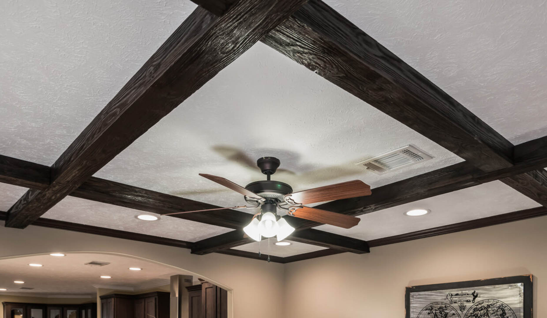 Ceiling with dark wooden beams in a grid pattern, featuring a central ceiling fan with wooden blades and lit bulbs. The room is softly lit and cozy.