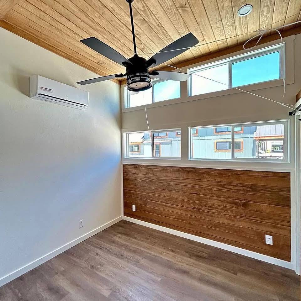 A small, bright room with light wood floors and ceiling. Large square windows let in natural light. A ceiling fan and air conditioner are visible. Cozy atmosphere.