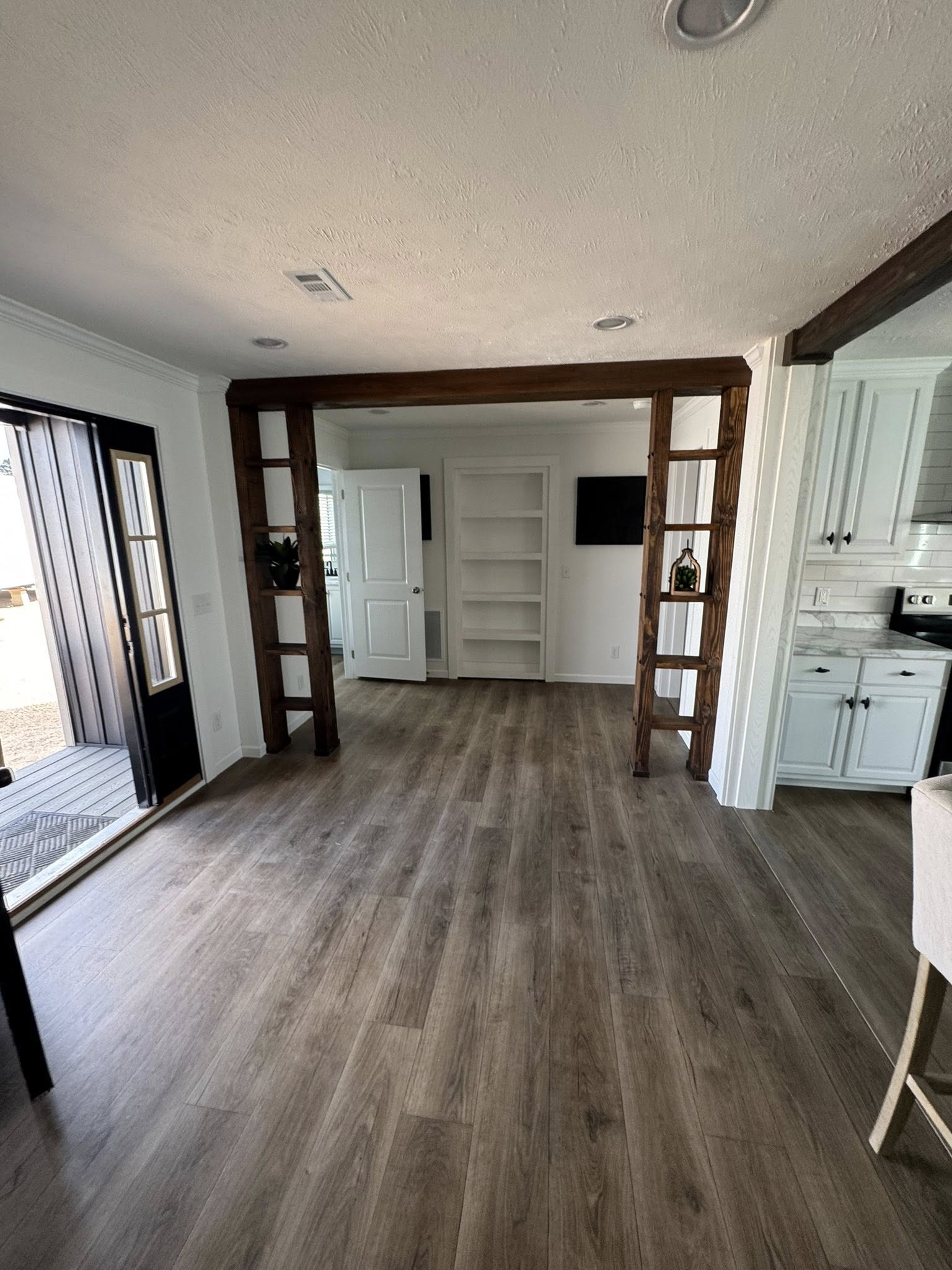Bright room with wooden floor, white walls, and kitchen view on the right. Open shelves frame a doorway to a room with built-in shelves and a black TV.