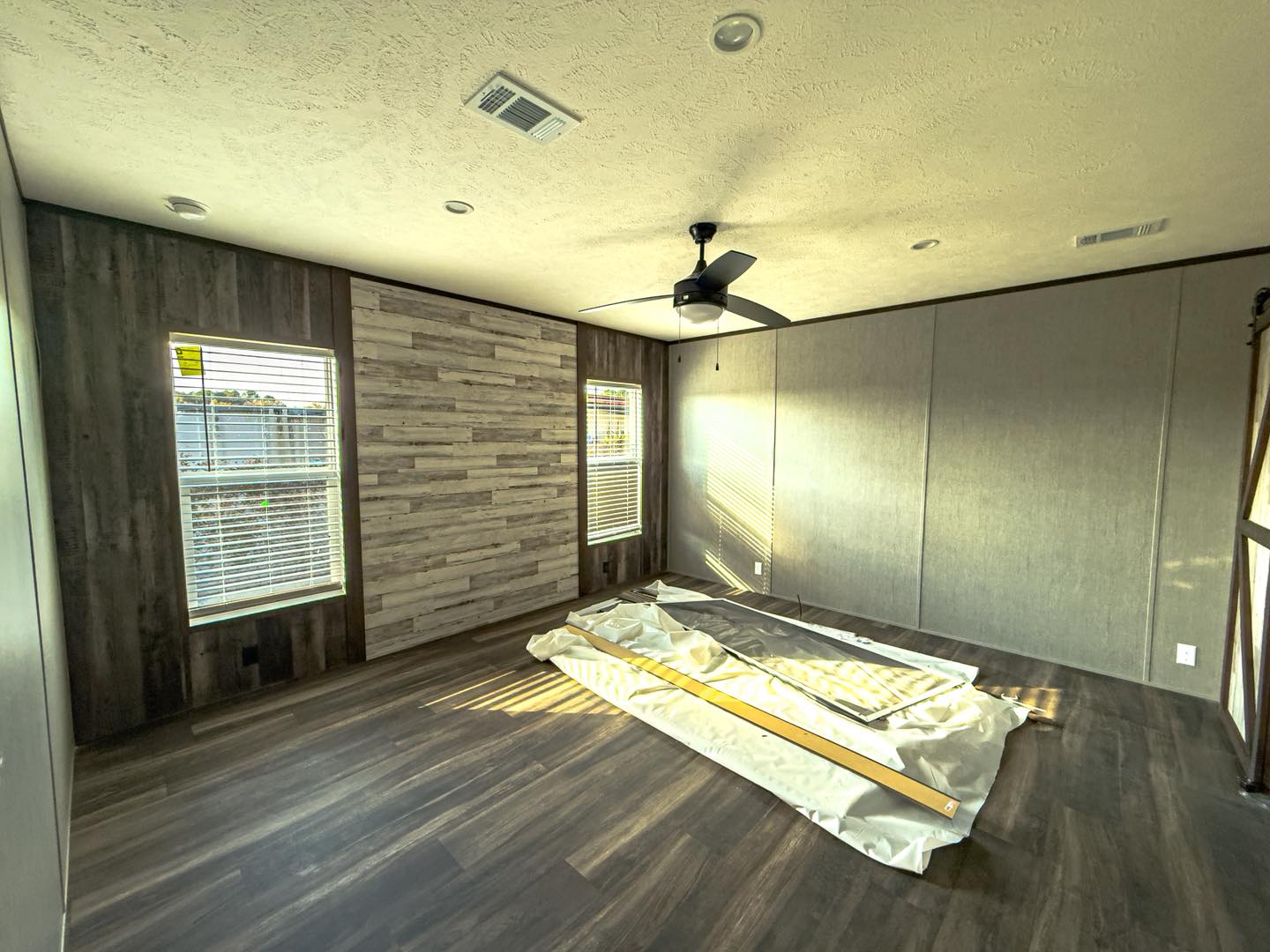 Empty room with wood panel walls and two windows. Sunlight filters in, casting warm shadows on the dark wood floor. Ceiling fan centered above.