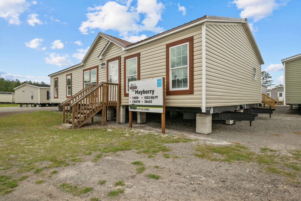 A beige modular home on a lot with a sign reading "Mayberry" indicating 3 beds, 2 baths, and 1352 sq ft. The setting is calm and sunny.