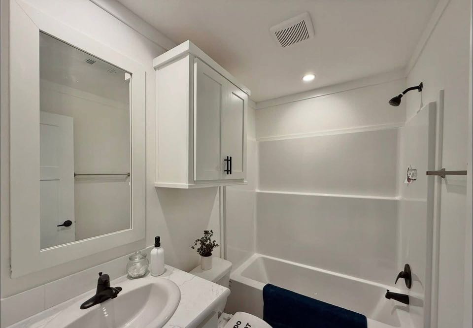 A small, modern bathroom with white walls and fixtures. Features a bathtub with black showerhead, a vanity with a sink, and a mirrored cabinet above.