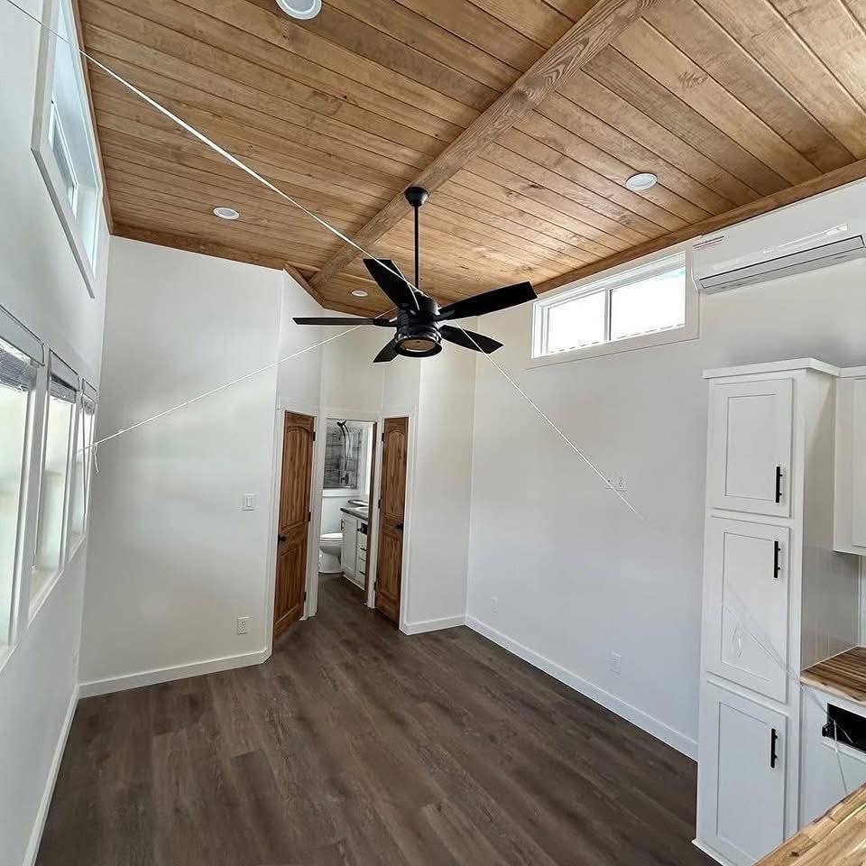 Compact room with a wooden ceiling and dark fan. White walls and cabinets create a bright space. Wooden floors lead to a small bathroom with double doors. Minimalist and cozy ambiance.