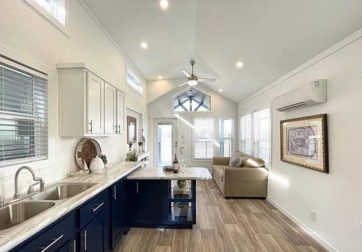 Bright, modern living space with white walls, high ceiling, and wood flooring. Kitchen on left; cozy seating area with sofa and wall art on right. Airy and inviting.