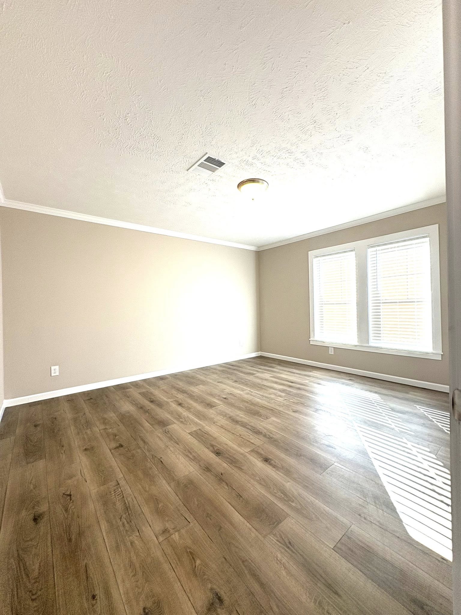 Bright, empty room with beige walls, large window with blinds, and wood laminate flooring. Sunlight casts soft shadows, creating a warm, inviting ambiance.