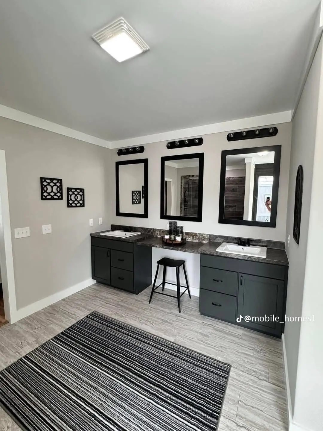 Modern bathroom with two sinks, black-framed mirrors, decorative wall art, and a central stool. Neutral tones and a striped rug create a cozy feel.