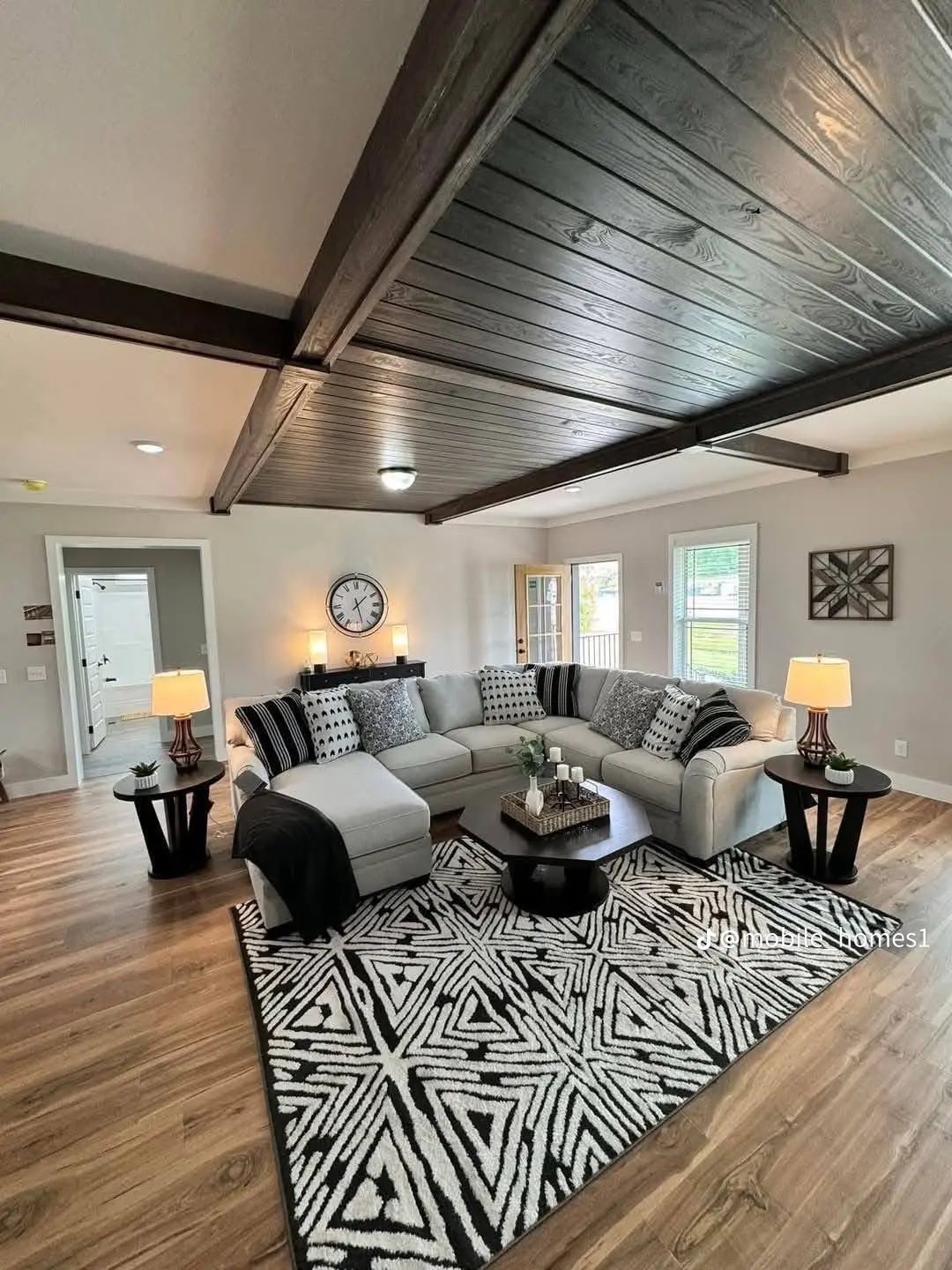 Spacious living room with a gray sectional sofa, patterned cushions, and a geometric rug. Dark wood beam ceiling, lamps on side tables, and a wall clock.