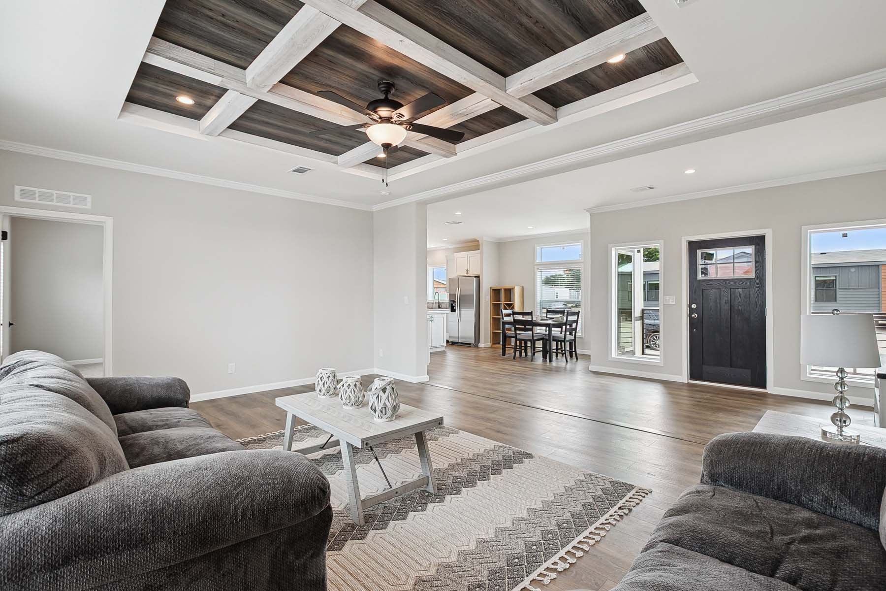Spacious living room with gray sofas, a patterned rug, and a white coffee table. Modern ceiling design, view of dining area and kitchen. Bright, airy feel.