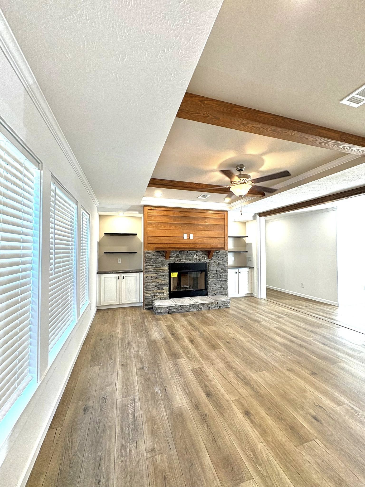 A spacious living room features a stone fireplace and wooden mantle, flanked by shelves and cabinets. The room has light wood flooring, ceiling beams, and a fan. Large windows with blinds allow natural light to fill the space.
