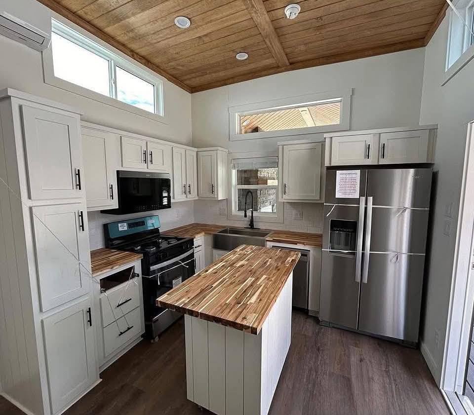 A modern kitchen with white cabinets, wooden countertops, and a central island. Stainless steel appliances include a refrigerator and a gas stove. Ample natural light from windows enhances the warm, inviting space.