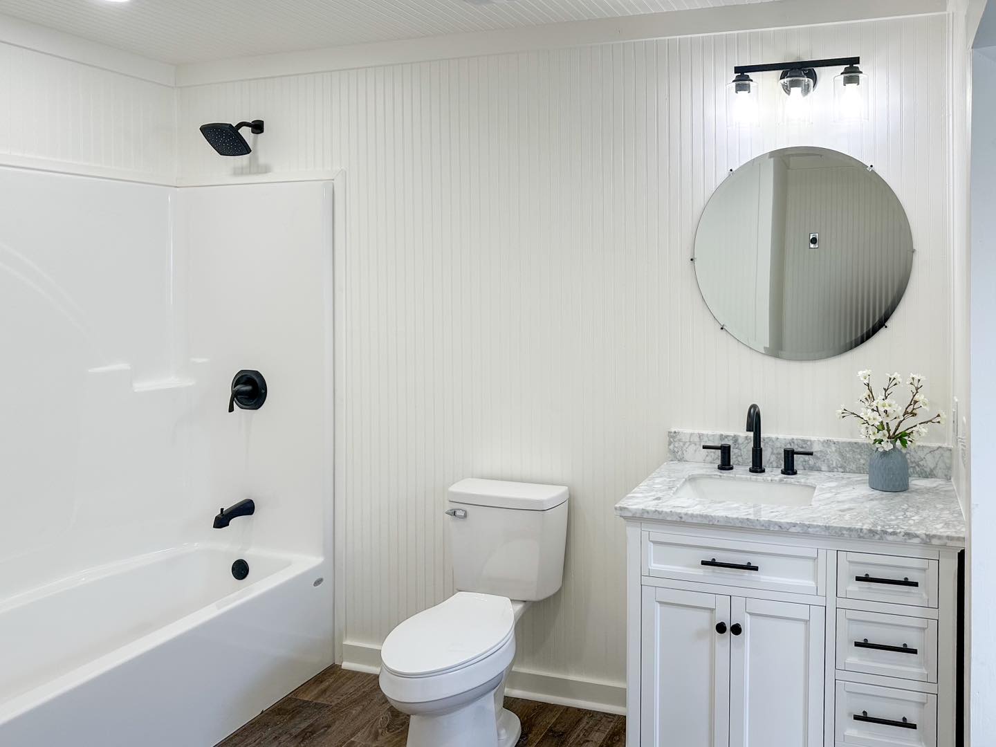 Modern bathroom with white walls and a bathtub on the left. A round mirror and vanity with marble top are on the right. Overall, clean and minimalist.