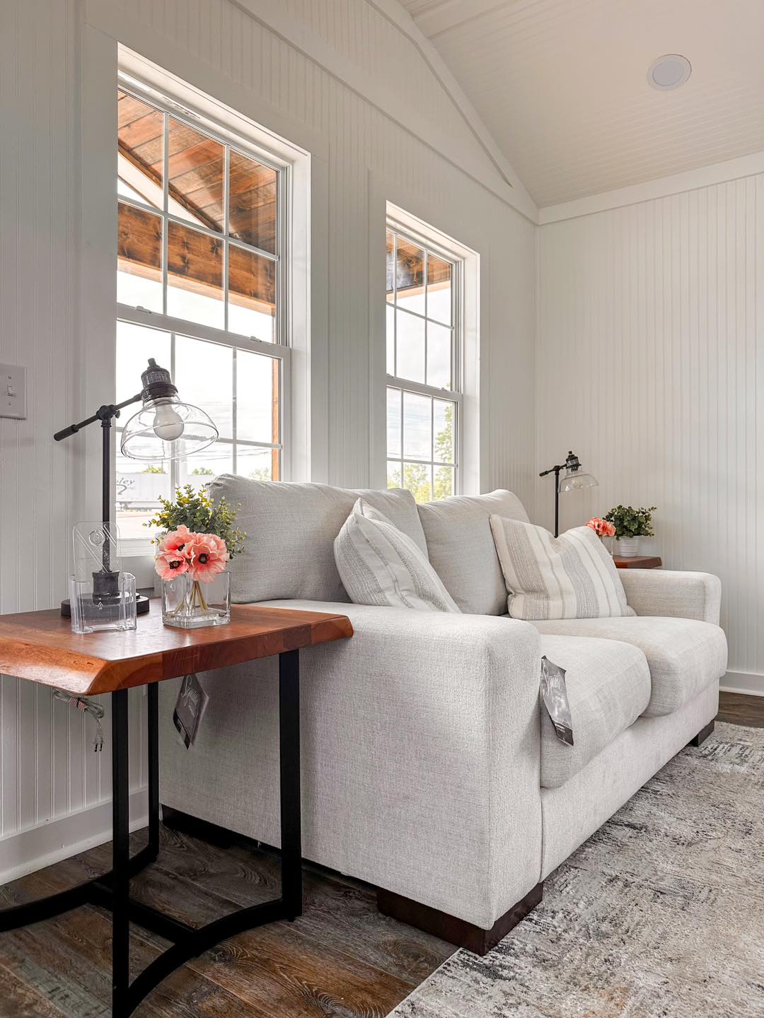 Cozy living room with a plush beige sofa, striped cushions, and wooden side tables. Soft pink flowers and lamps add warmth near tall windows.