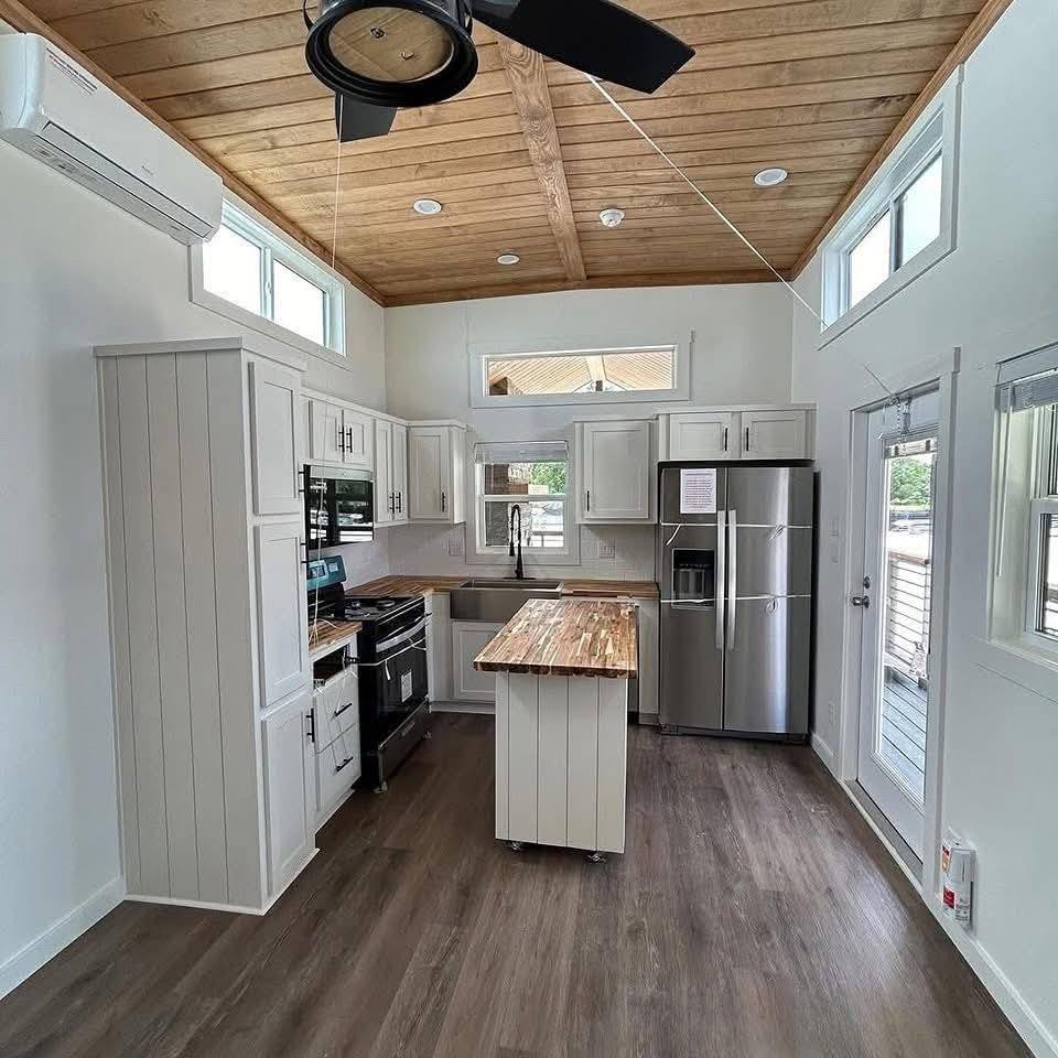 Modern kitchen with wooden ceiling and floor, featuring white cabinets, stainless steel appliances, a central island, and large windows for natural light.