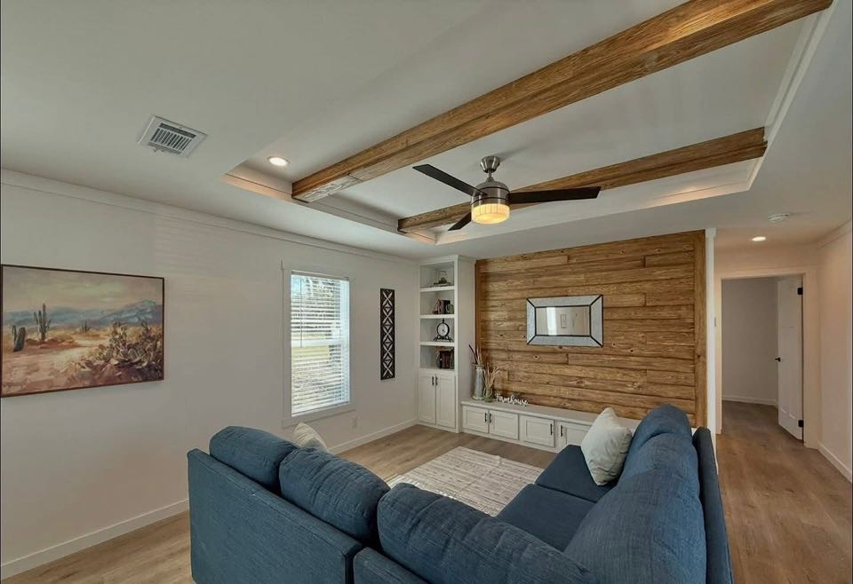 A cozy living room with a wooden accent wall, blue sectional sofa, large ceiling fan, and natural wood beams. Sunlit with a serene, inviting atmosphere.