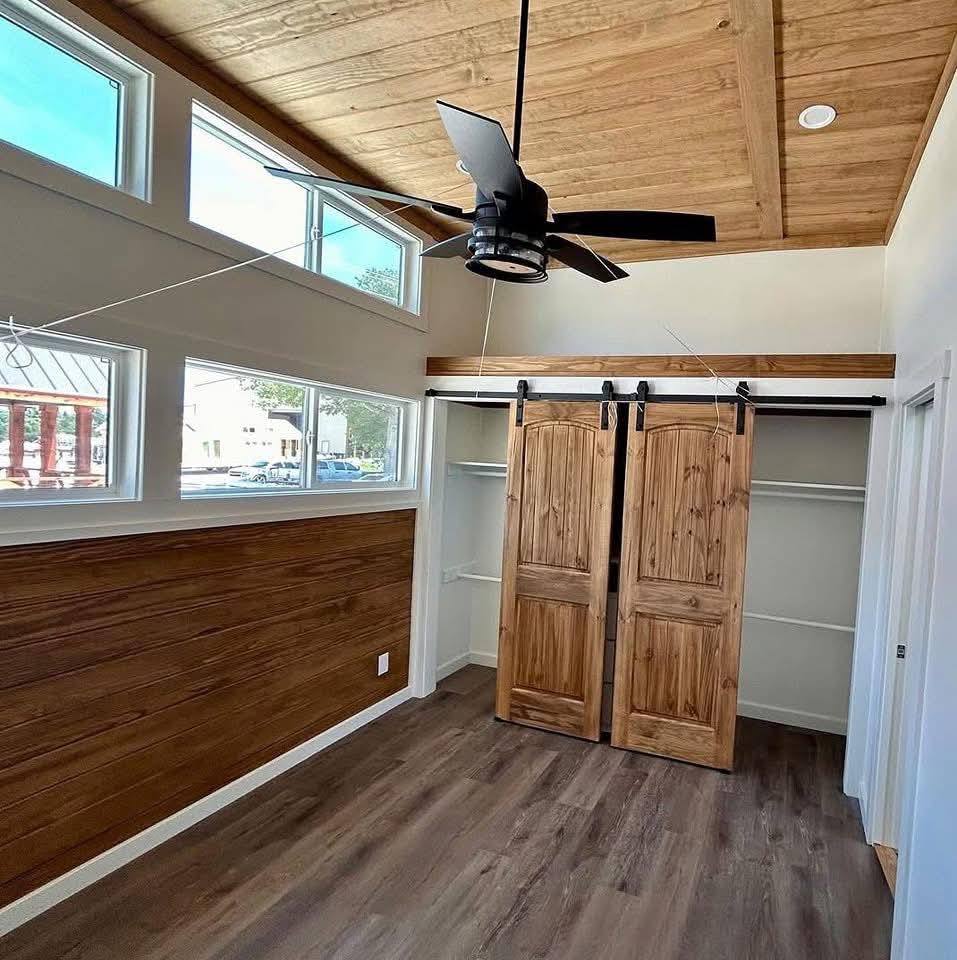Cozy room with wood paneling and ceiling, featuring tall windows, wooden sliding closet doors, and a modern black ceiling fan, creating a warm ambiance.