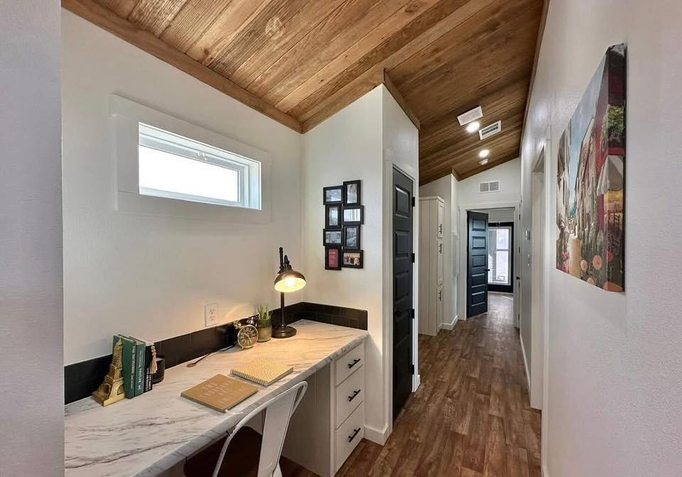 Cozy hallway with a wooden ceiling and floor, featuring a corner desk with a lamp, books, and plants. Black doors contrast with white walls.