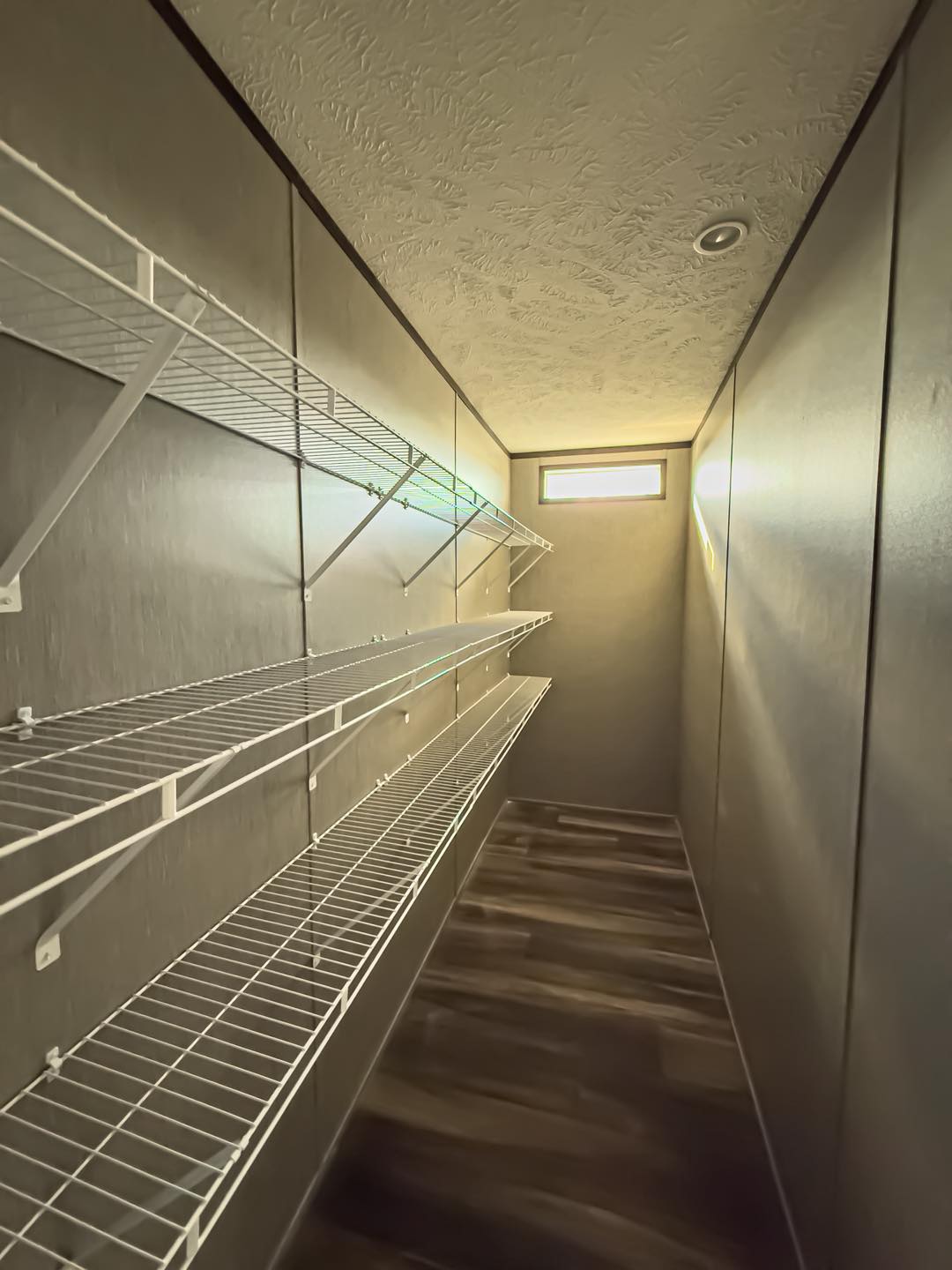 Narrow room with wire shelves on the left wall, beige walls, and brown wood floor. Small window at the end provides natural light, creating a calm atmosphere.