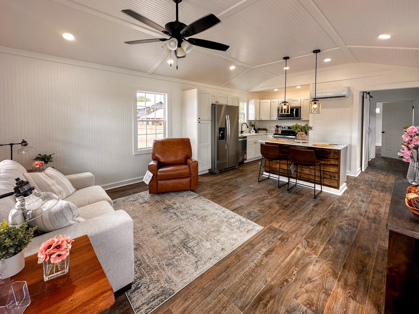 Bright open-concept living room and kitchen with wood floors. Features a white sofa, leather chair, rustic kitchen island, and modern appliances.