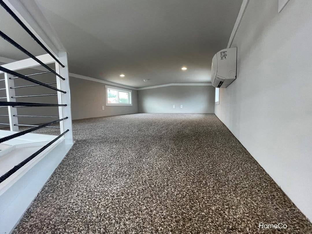Cozy carpeted attic space with white walls and small windows. Recessed lighting brightens the room. An AC unit is mounted on the right wall.