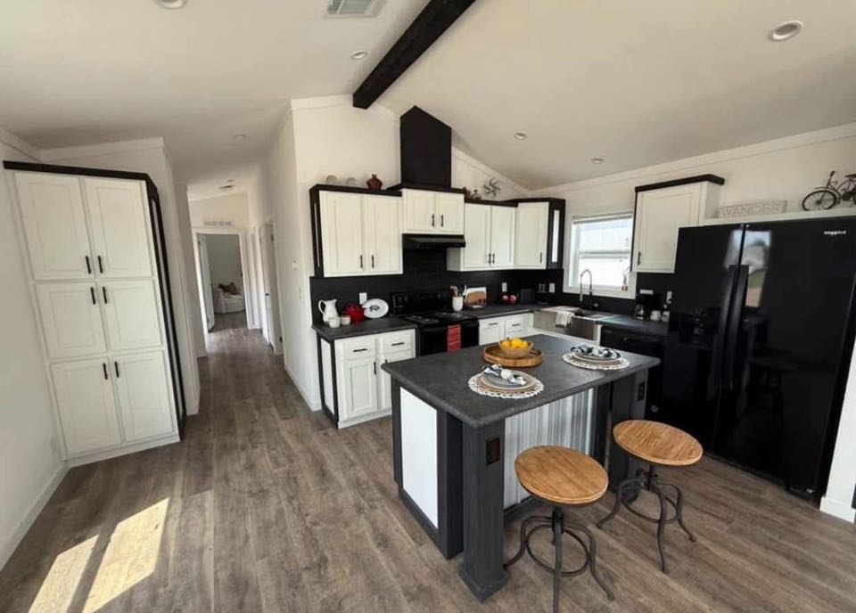 Modern kitchen with a monochrome color scheme features white cabinets, black countertops, and a black refrigerator. The island has wooden bar stools and a fruit bowl. Warm lighting and wooden floors create a cozy atmosphere.