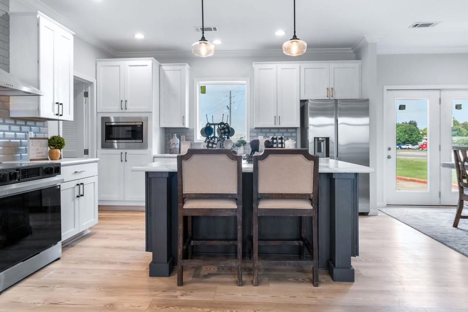 Modern kitchen with white cabinets, a central island with two chairs, stainless steel appliances, wooden floor, and pendant lights. Bright and inviting atmosphere.