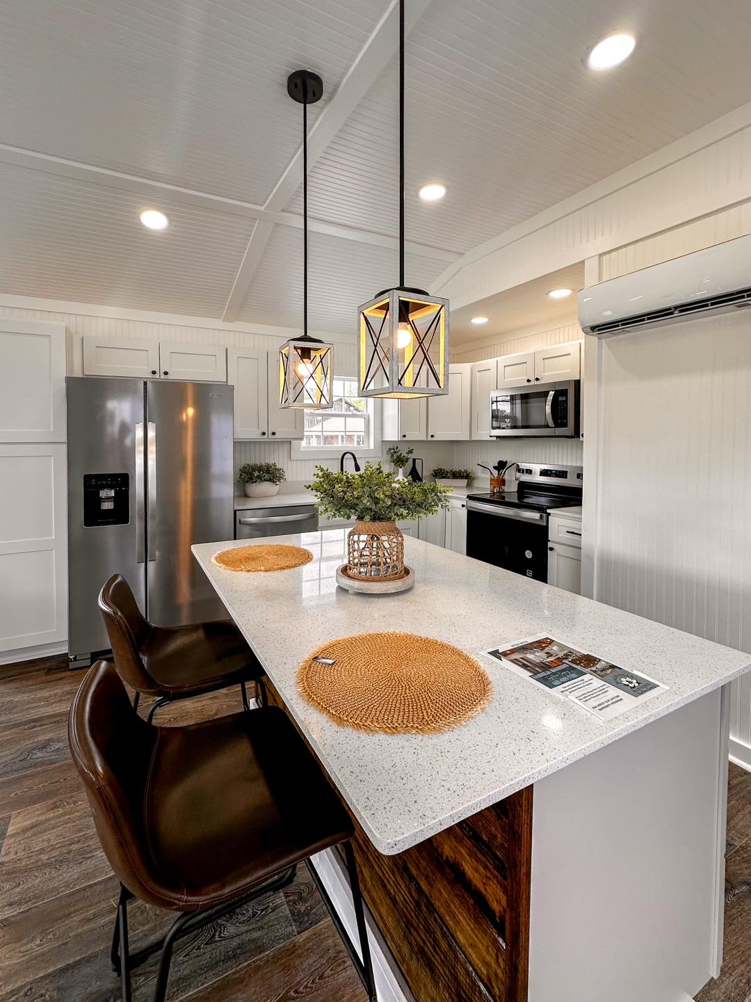 Modern kitchen with white cabinets, island with two brown chairs, wicker placemats, and pendant lights. Stainless steel appliances add sleekness.