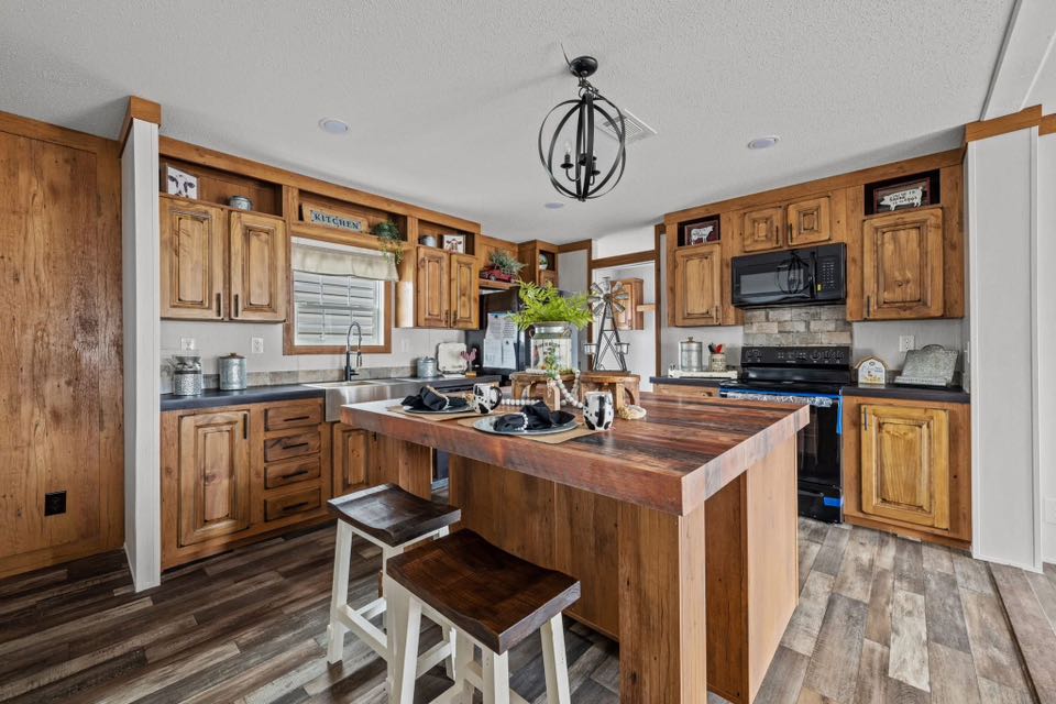 Rustic kitchen with wooden cabinets and flooring. A central island features place settings and stools. Black appliances add contrast, creating a cozy vibe.