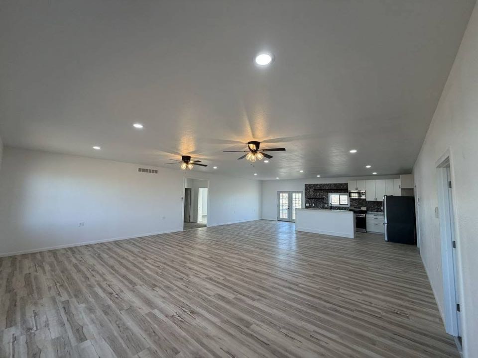 Spacious, modern open-concept living area with light wood flooring, ceiling fans, and recessed lighting. Kitchen in background, featuring dark backsplash.