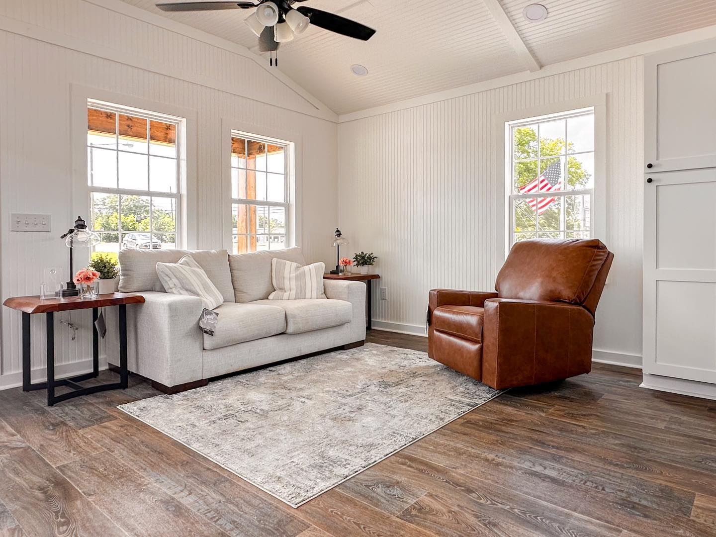 A cozy living room with a beige sofa, striped pillows, and a brown leather recliner on a patterned rug. Sunlight filters through white-framed windows.