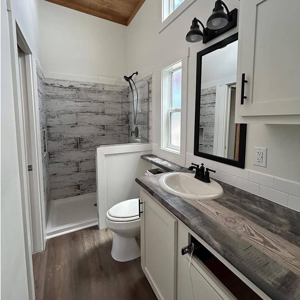 Small, modern bathroom with rustic wood accents. Features a gray, wood-paneled shower, white fixtures, dark wood countertop, and minimalist decor.