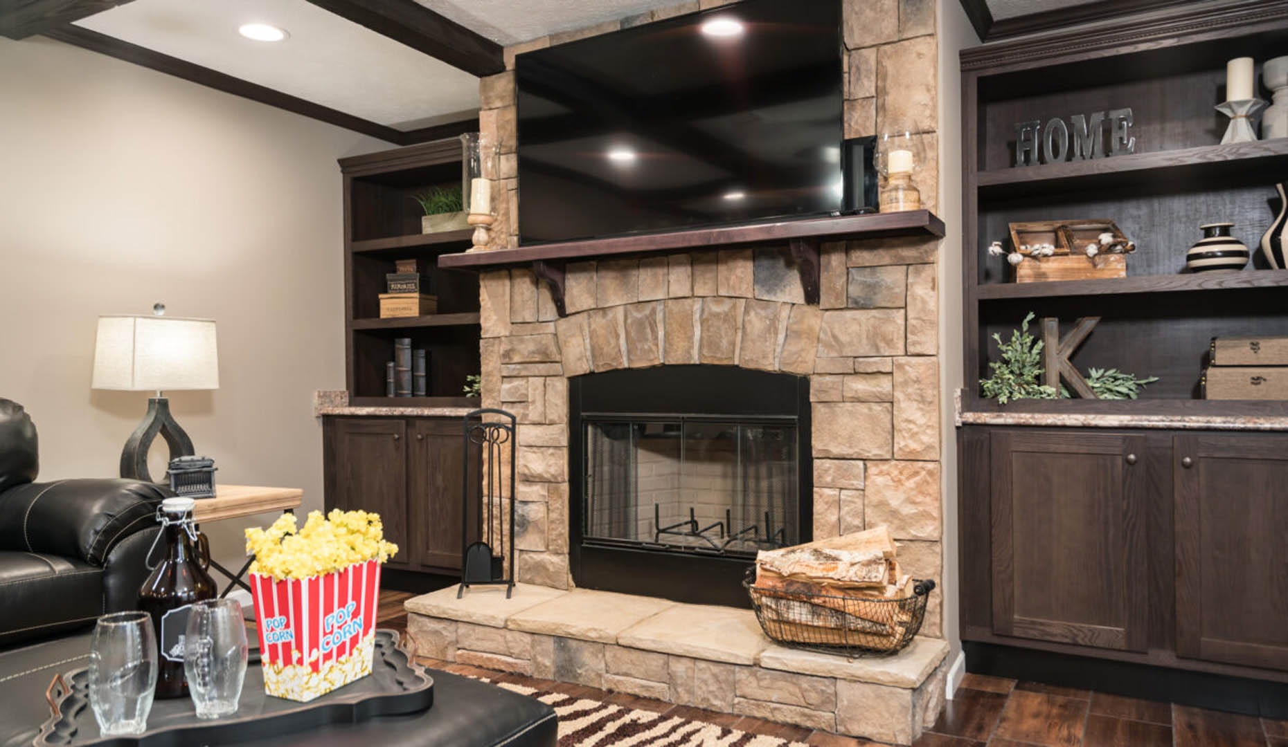 Cozy living room with stone fireplace and large TV. Dark wood shelves hold decor. A lamp and a tray with popcorn, bottles, and glasses on a leather ottoman add warmth.