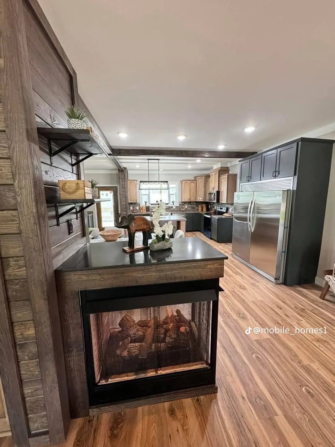 Modern kitchen with wood floors, a central island, and sleek stainless steel fridge. Cozy electric fireplace with decorative plant on top sits nearby.