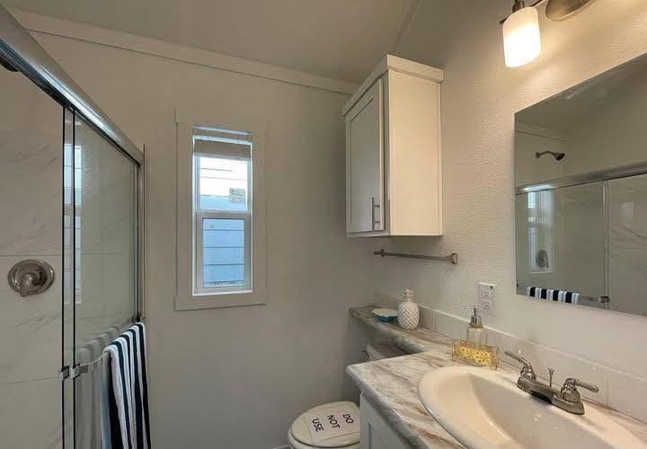 Small, bright bathroom with a shower, striped towels, and marble countertop. A wall cabinet and warm lighting create a clean, inviting atmosphere.