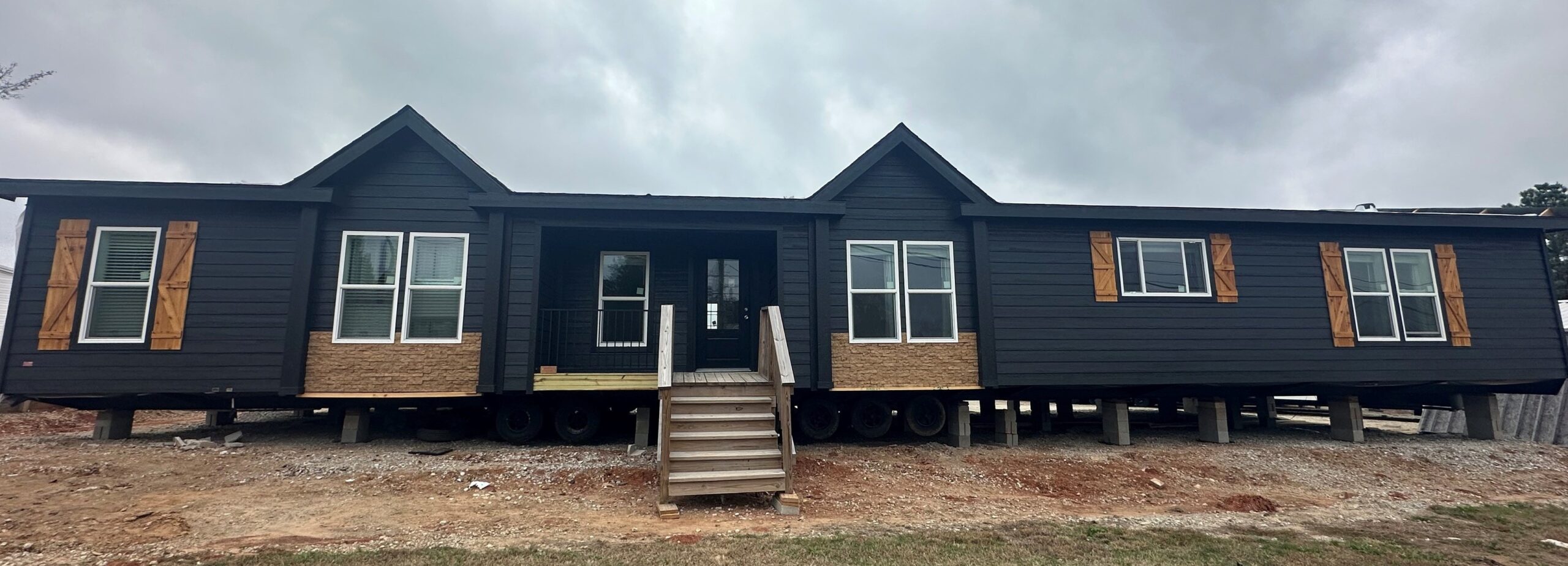 A large, dark gray modular home on wheels sits elevated on a dirt lot. It features three sections with white windows and rustic wooden shutters.